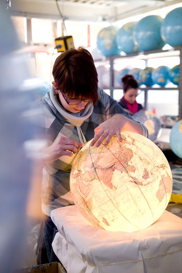 Person in Kapuzenpullover arbeitet an beleuchtetem Globus in Werkstatt mit weiteren Globen im Hintergrund.